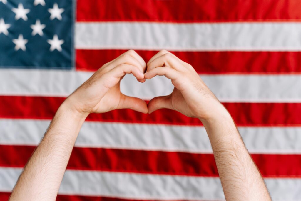 Hands forming a heart shape in front of the American flag, symbolizing love and patriotism.